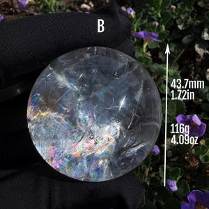 Peut inclure: Une boule de cristal sphérique et transparente marquée de la lettre B, présentant des fractures internes qui créent des réfractions lumineuses arc-en-ciel. La sphère mesure 43,7 mm de diamètre et pèse 116 g, selon le texte sur l'image.