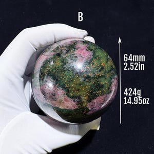 Könnte beinhalten: Eine polierte Edelstein-Kugel, hauptsächlich grün mit rosa und goldenen Einschlüssen. Die Kugel hat einen Durchmesser von 64 mm und wiegt 424 g. Der Buchstabe "B" ist oben sichtbar. Die Kugel wird von einer behandschuhten Hand gehalten.