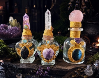 Könnte beinhalten: Drei klare Glasflaschen mit goldfarbenen Verzierungen und Edelstein-Verschlüssen. Zwei Flaschen haben spitze Kristallkristalle, während die dritte eine runde Rosenquarzkugel besitzt. Jede Flasche ist mit eingebetteten Steinen und Blattmotiven verziert.