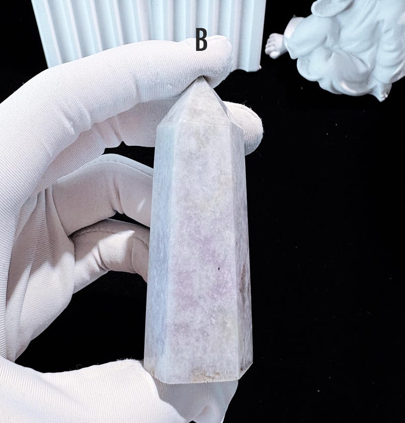 Puede incluir: Un obelisco de cristal morado claro con un dise&ntilde;o facetado. El cristal se sostiene en una mano enguantada sobre un fondo negro. El obelisco tiene una parte superior puntiaguda y una base rectangular.