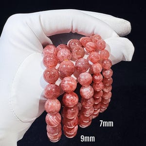 May include: Three bracelets of strawberry quartz beads in 7mm, 9mm, and 10mm sizes. The beads are a translucent pink with red inclusions. The bracelets are held against a dark background.