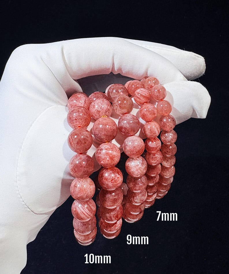 May include: Three bracelets of strawberry quartz beads in 7mm, 9mm, and 10mm sizes. The beads are a translucent pink with red inclusions. The bracelets are held against a dark background.