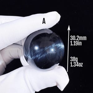 May include: A polished blue tiger's eye sphere labelled A, measuring 30.2mm or 1.19 inches in diameter and weighing 38g or 1.34 ounces. The stone features a dark blue base with chatoyant light blue bands that create a shimmering, reflective effect.