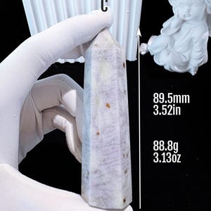 Puede incluir: Un obelisco de cristal morado claro es sostenido por una mano enguantada. El obelisco mide 89,5 mm de alto y pesa 88,8 g. El cristal es de color morado claro y blanco con motas marrones. Una estatua blanca de un &aacute;ngel est&aacute; en el fondo.