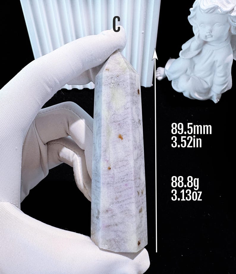 Puede incluir: Un obelisco de cristal morado claro es sostenido por una mano enguantada. El obelisco mide 89,5 mm de alto y pesa 88,8 g. El cristal es de color morado claro y blanco con motas marrones. Una estatua blanca de un &aacute;ngel est&aacute; en el fondo.