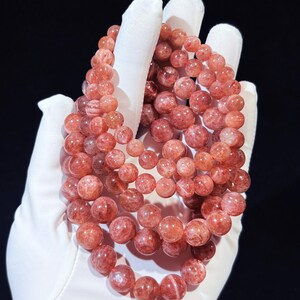May include: Several bracelets made of reddish-orange gemstone beads. The beads are round and held in a white gloved hand against a black backdrop. The beads have a translucent appearance with varying shades of colour.