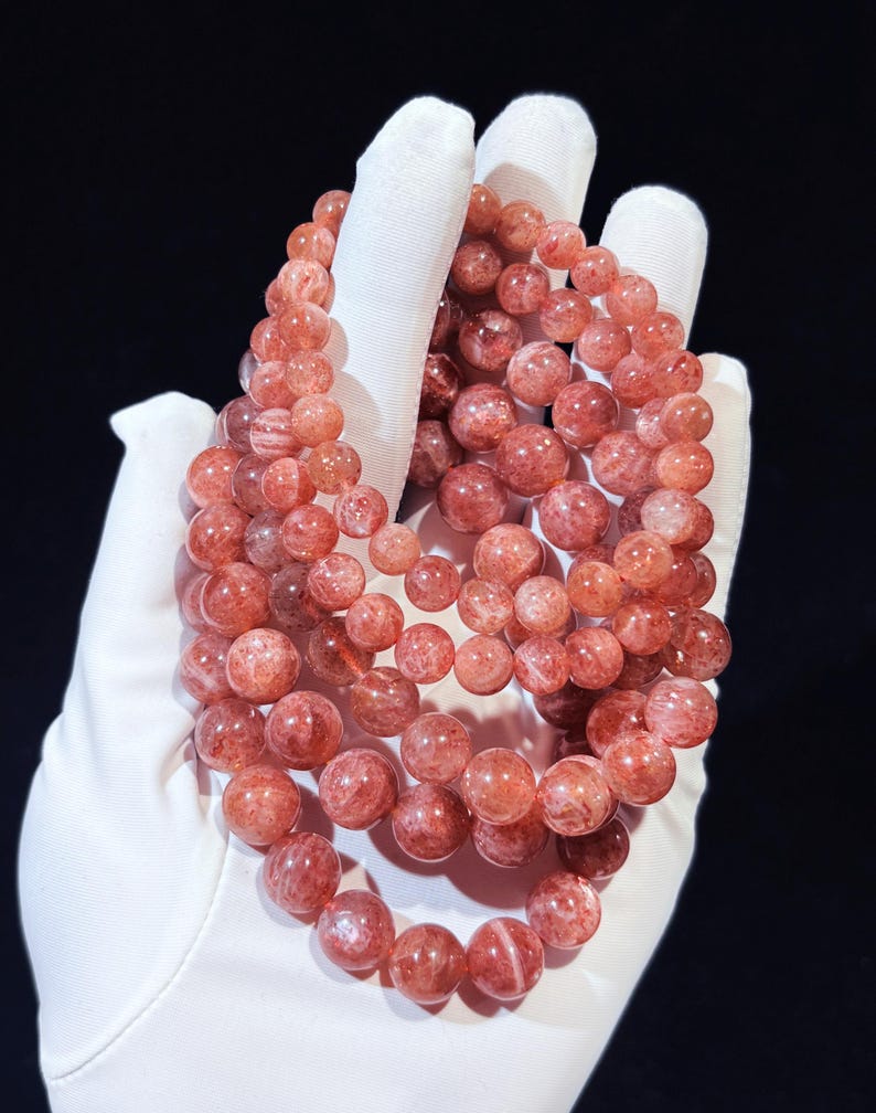 May include: Several bracelets made of reddish-orange gemstone beads. The beads are round and held in a white gloved hand against a black backdrop. The beads have a translucent appearance with varying shades of colour.
