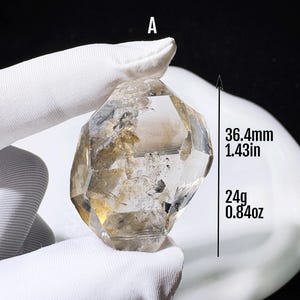 Könnte beinhalten: Ein klarer, facettierter Quarzkristall mit der Kennzeichnung A, der interne Einschlüsse und goldbraune Farbtöne aufweist. Der Kristall ist 36,4 mm hoch und wiegt 24 g, wie durch den Text und die Pfeilgrafik neben dem Stein angegeben.