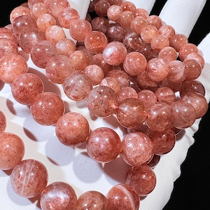May include: Close-up of several strands of round, reddish-orange gemstone beads. The beads have a speckled appearance and are strung together, likely for bracelets or necklaces. The beads are held in a white gloved hand.