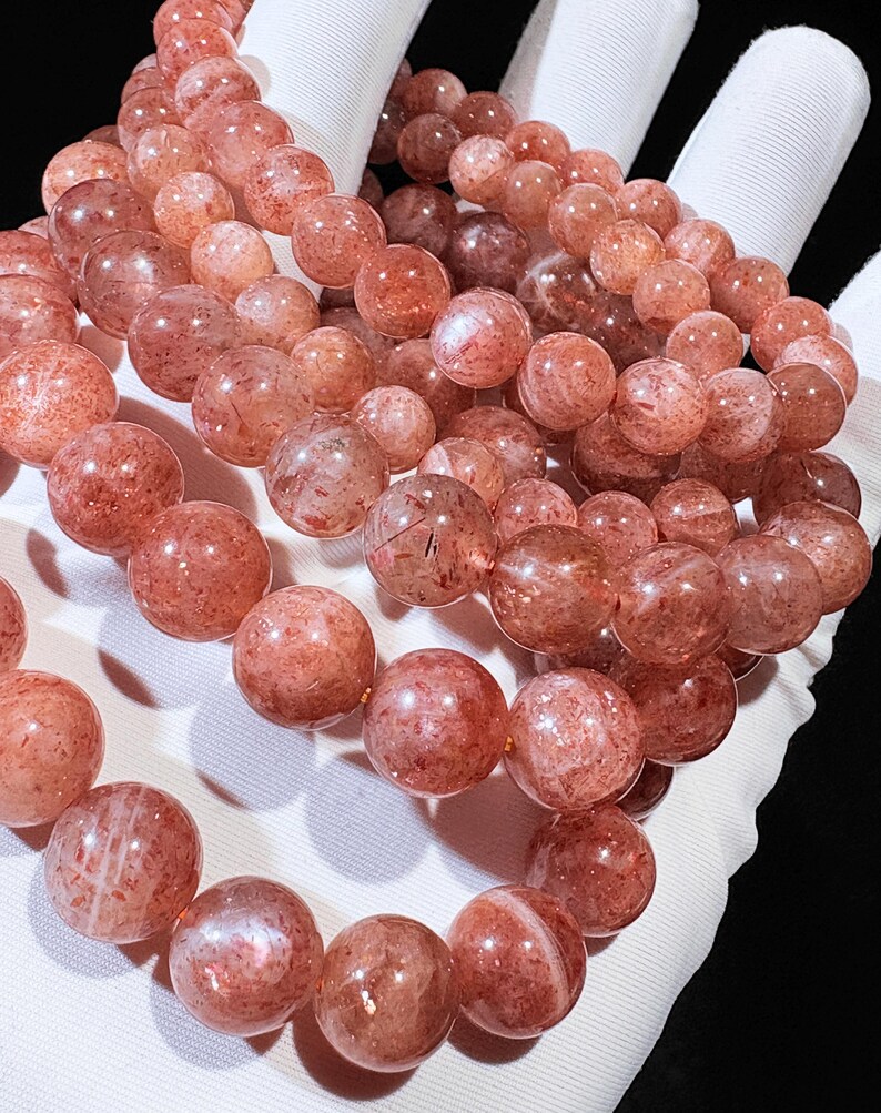 May include: Close-up of several strands of round, reddish-orange gemstone beads. The beads have a speckled appearance and are strung together, likely for bracelets or necklaces. The beads are held in a white gloved hand.