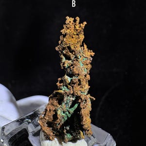 Puede incluir: Una muestra de mineral de cobre con una superficie rugosa y texturizada. El mineral muestra una mezcla de tonos marrones y verdes, con la letra "B" visible. La muestra est&aacute; montada sobre una base transparente similar al vidrio.