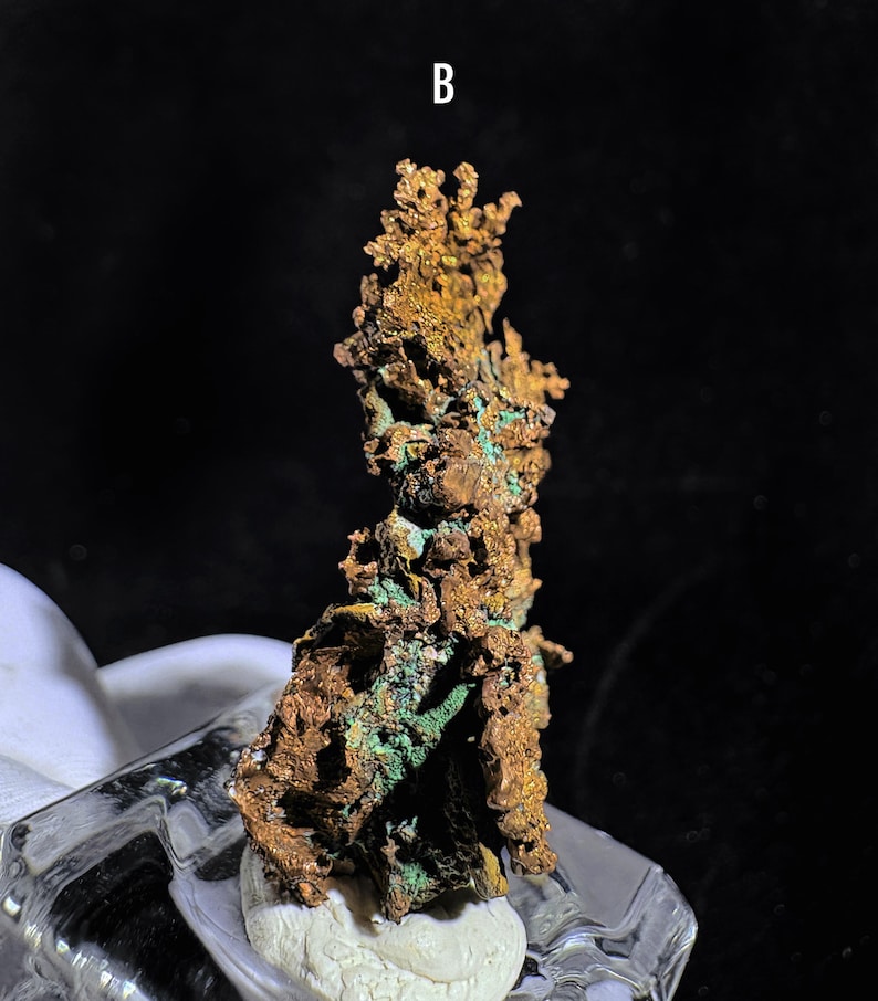 Puede incluir: Una muestra de mineral de cobre con una superficie rugosa y texturizada. El mineral muestra una mezcla de tonos marrones y verdes, con la letra "B" visible. La muestra est&aacute; montada sobre una base transparente similar al vidrio.