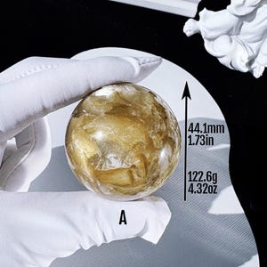 May include: A golden-yellow crystal sphere, 44.1mm in diameter and weighing 122.6g, is held by a gloved hand. The sphere has a textured, translucent appearance. The letter "A" is printed on the surface.