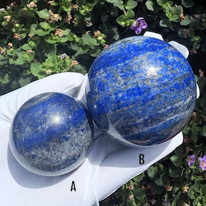 May include: Two polished lapis lazuli spheres labelled A and B. The spheres are deep blue with visible gold-coloured pyrite inclusions and white mineral veining. Sphere A is smaller than sphere B, and both are resting on a white fabric surface.