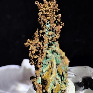 Puede incluir: Primer plano de un esp&eacute;cimen mineral de cobre natural. El mineral tiene una superficie rugosa y texturizada con una mezcla de tonos dorados, marrones y verdes. La letra "B" es visible en la esquina superior izquierda. El mineral se exhibe sobre una superficie blanca.