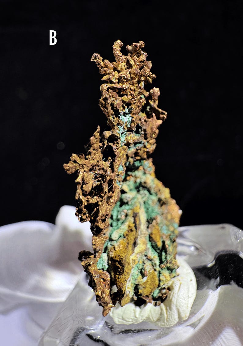 Puede incluir: Primer plano de un esp&eacute;cimen mineral de cobre natural. El mineral tiene una superficie rugosa y texturizada con una mezcla de tonos dorados, marrones y verdes. La letra "B" es visible en la esquina superior izquierda. El mineral se exhibe sobre una superficie blanca.