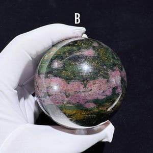 Puede incluir: Una esfera de piedra preciosa pulida con una mezcla de colores verdes, rosas y dorados. La esfera es sostenida por una mano enguantada, con la letra "B" en el fondo. La superficie de la esfera es reflectante.