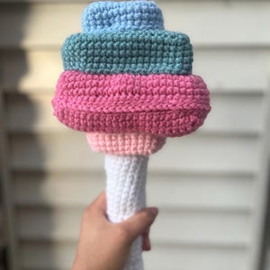 May include: A handmade crochet ice cream cone toy. The toy features a white handle and a colorful ice cream stack in shades of pink, green, and blue. The crochet stitches are visible, giving it a textured appearance. The toy is designed to resemble a soft-serve ice cream cone.
