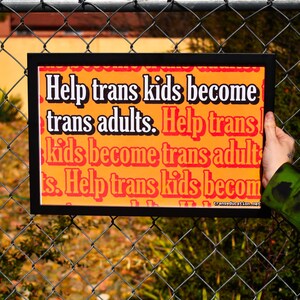 May include: A black framed sign with an orange background and white text that reads "Help trans kids become trans adults. Help trans kids become trans adults. Help trans kids become trans adults." The sign is hanging on a chain link fence.