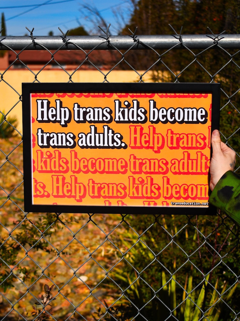May include: A black framed sign with an orange background and white text that reads "Help trans kids become trans adults. Help trans kids become trans adults. Help trans kids become trans adults." The sign is hanging on a chain link fence.
