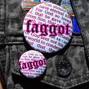 Op de afbeelding: Close-up van een donker denim jack versierd met verschillende ronde knopen. De knopen tonen het woord "faggot" in roze gotische letters, omringd door religieuze tekst. Extra patches en een knoop zijn ook zichtbaar.