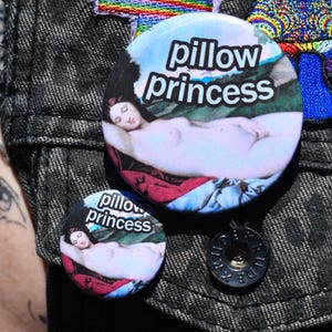 May include: Two round buttons with the text "pillow princess" and an image of a reclining figure. The larger button is approximately 1.5 inches in diameter, and the smaller one is about 1 inch. The buttons are on a dark denim jacket.