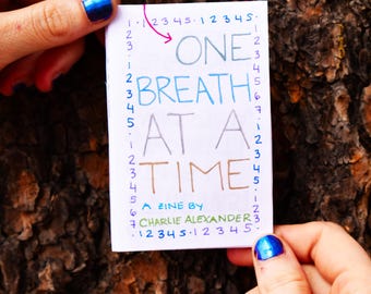 Può includere: Un biglietto bianco con testo scritto a mano in blu, verde e rosso che dice "One Breath At A Time. A Zine by Charlie Alexander." Il biglietto è tenuto da due mani con smalto per unghie blu.