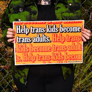 May include: A person holding a sign that reads "Help trans kids become trans adults. Help trans kids become trans adults. Help trans kids become trans adults." The sign is orange with black text and a website address at the bottom.
