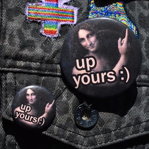 May include: Two round black buttons with the text "up yours :) " and an image of a woman pointing up. The buttons are on a dark denim jacket, along with other colorful patches.