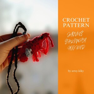 Puede incluir: Un camarón rojo tejido a crochet con ojos negros y una cáscara azul y blanca. El camarón está siendo sostenido por una mano y está unido a un cordón negro. La imagen tiene el texto "CROCHET PATTERN HOMES SHRIMPIFY HOOKED by artsy.kiky" en texto blanco sobre un fondo naranja.