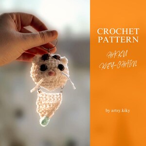 May include: White crocheted cat keychain with black eyes and a pink nose. The keychain is attached to a silver ring. The image is against a blurred background.