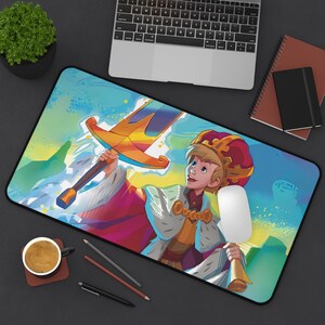 May include: A colorful desk mat featuring a cartoon illustration of a young king holding a sword, wearing a red and gold crown and a blue and gold robe. The background is a blue and green landscape with clouds and mountains.