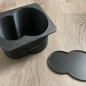 May include: Black plastic cup holder with two compartments and a removable lid. The lid is shaped like a figure eight.