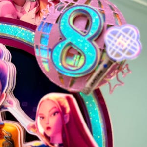 May include: A colorful cake topper featuring a large teal number 8 with glitter accents, set against a pink disco ball design. The topper includes cartoon figures and a microphone, suggesting a birthday or celebration theme.