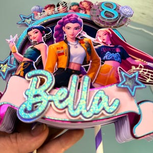 May include: A colorful cake topper featuring three animated female figures with purple, blue, and pink hair. The topper includes the name "Bella" in glittery blue letters, a number "8", and musical notes. The figures are wearing stylish outfits.