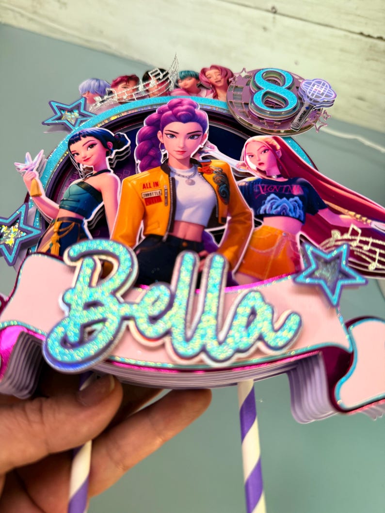May include: A colorful cake topper featuring three animated female figures with purple, blue, and pink hair. The topper includes the name "Bella" in glittery blue letters, a number "8", and musical notes. The figures are wearing stylish outfits.