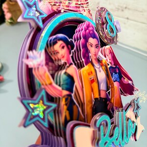 May include: A multi-layered cake topper featuring anime-style characters in vibrant colors. The design includes a teal-colored "Bella" script, stars, and a number "8". The topper has a purple, teal, and yellow color scheme.