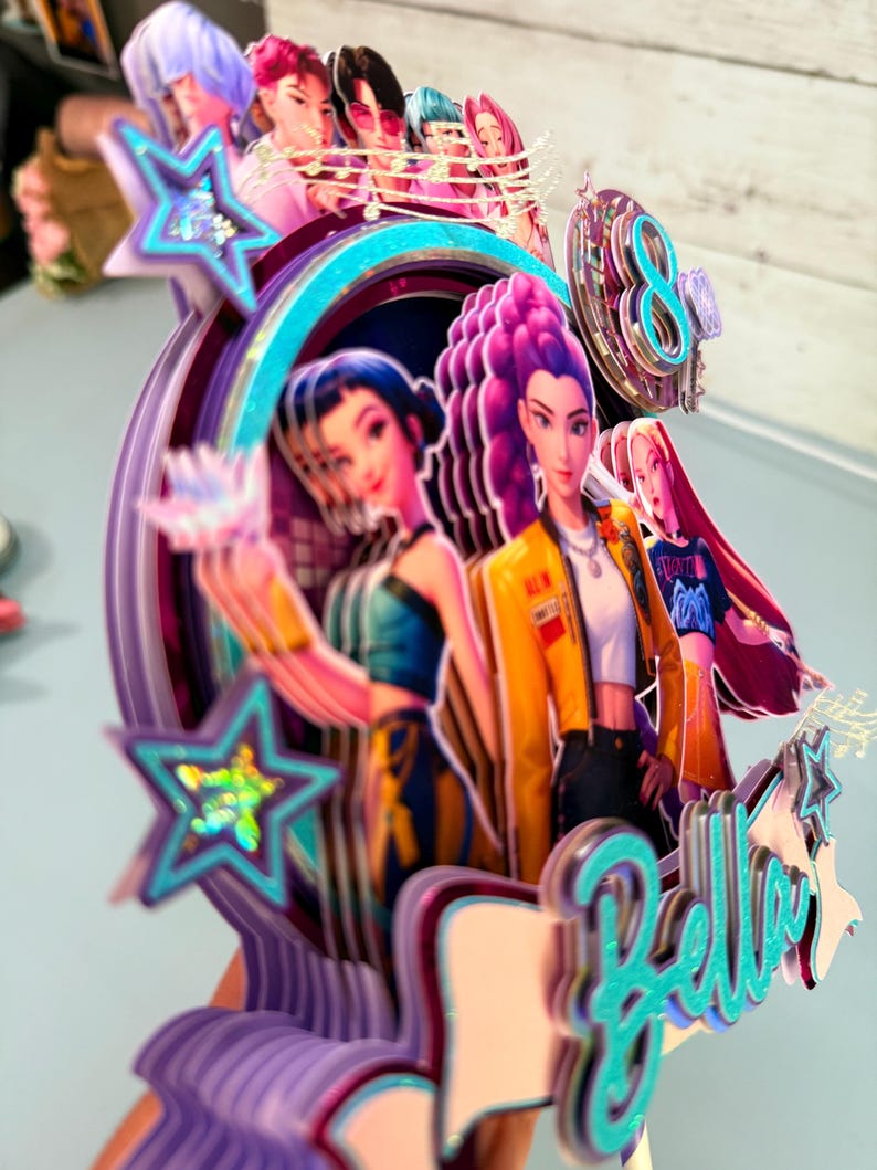 May include: A multi-layered cake topper featuring anime-style characters in vibrant colors. The design includes a teal-colored "Bella" script, stars, and a number "8". The topper has a purple, teal, and yellow color scheme.
