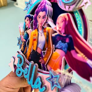 May include: A multi-layered cake topper featuring animated female characters in vibrant colors. The topper includes the name "Bella" in blue script, surrounded by stars and a circular frame. The characters are depicted in various outfits and hairstyles.