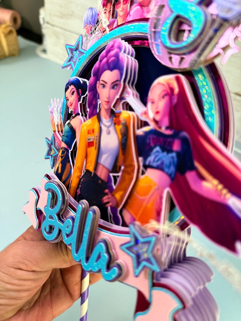 May include: A multi-layered cake topper featuring animated female characters in vibrant colors. The topper includes the name "Bella" in blue script, surrounded by stars and a circular frame. The characters are depicted in various outfits and hairstyles.