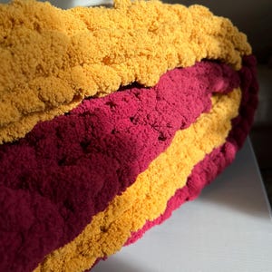 May include: A handmade, chunky knit blanket in a sandwich-like design. The blanket features alternating layers of golden yellow and burgundy red yarn. The thick, textured yarn creates a cozy and inviting appearance, perfect for snuggling.