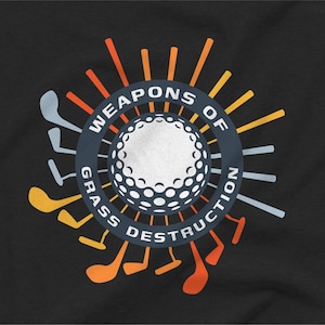 May include: Black t-shirt with a golf-themed graphic. The design features a golf ball at the center, surrounded by the text "WEAPONS OF GRASS DESTRUCTION" and golf clubs in various colors, including orange, yellow, and gray.