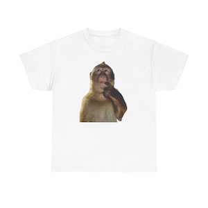 May include: White t-shirt featuring a brown monkey with its finger in its mouth. The monkey has brown fur and is set against a white background. The t-shirt is a classic crew neck style.