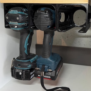 Puede incluir: Dos taladros inalámbricos, uno azul y negro y otro negro y rojo, están colgados en un soporte de almacenamiento de pared. El taladro azul y negro tiene una batería negra y el taladro negro y rojo tiene una batería roja. Los taladros están colgados por sus clips de cinturón.