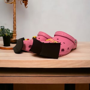 Puede incluir: Dos zuecos Crocs rosas con abalorios Jibbitz negros. Los zuecos están sobre una superficie de madera.