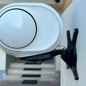 May include: White wireless speaker mounted on a black wall mount bracket. The speaker is attached to the wall with screws. The speaker has a round, white front with a black trim around the edge.