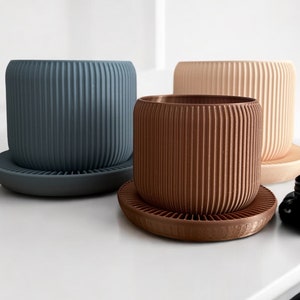 Puede incluir: Tres macetas de cerámica con platillos a juego. Las macetas son acanaladas y vienen en azul, marrón y beige.