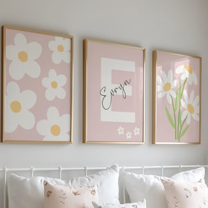 May include: Three framed wall art prints with gold frames hanging above a bed. The left print features white daisies on a pink background. The center print displays a large white letter E with the name Evryn in black script. The right print shows white flowers with green stems on a pink background.