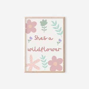 May include: A framed print with a light wood frame. The print features the text "Shes a wildflower" in a soft pink color, surrounded by pastel-colored floral illustrations in pink, green, and purple.