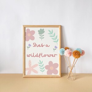 May include: A framed print with the text "She's a wildflower" and floral illustrations in pink, green, and purple. Beside it, a glass jar holds several pom-pom flowers in various pastel colors.
